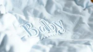 Close-up of soft embroidered lettering on a pale blue cotton baby blanket, showing delicate stitching detail, natural morning light, cozy nursery setting