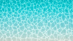 Overhead view of crystal-clear turquoise shallow water with white sand ripples visible beneath, gentle waves at shoreline, tropical beach setting